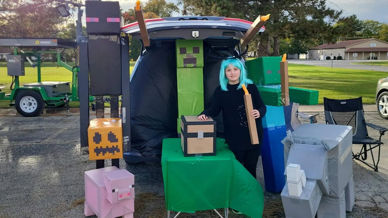 Photo of a car at trunk-or-treat event surrounded by a zombie, creeper, pig, enderman holding a jack-o-lantern, and torches with a Minecraft chest on a table that stores candy. 