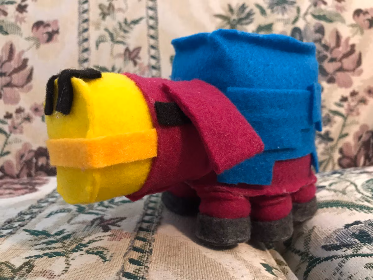 Photo of the sniffer, a Minecraft character, created out of felt. It has a rectangular body that is burgundy and teal atop four legs with a yellow nose