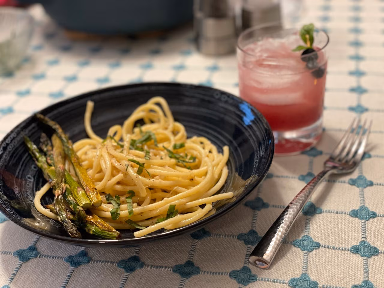 Photo of cooked noodles in a black bowl with a short glass filled with a pinkish beverage