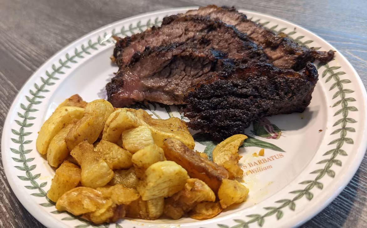 Photo of some sliced pieces of cooked brisket on a white plate with some roasted potatoes on the side