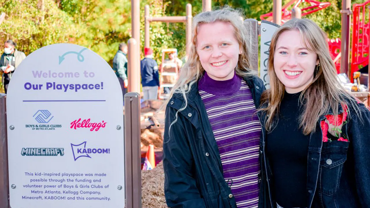 Morgan Sloane and another volunteer are next to a sign at a playground under construction which reads, Welcome to Our Playspace!